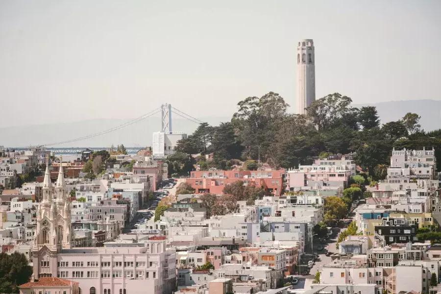 Se muestra la Torre Coit de San Francisco con el Puente de la Bahía al fondo y una colina cubierta de casas en primer plano.